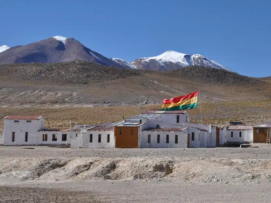 Bolivian tricolour flag flying above a remote building near Salar de Uyuni, with snow-capped mountains in the background