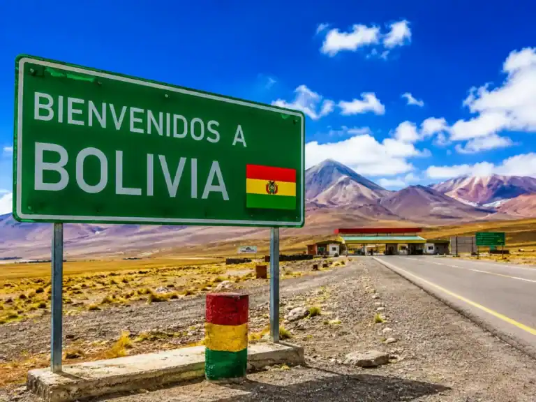 Roadside welcome sign at a Bolivia land border crossing with mountains, desert landscape, and immigration checkpoint in the background.