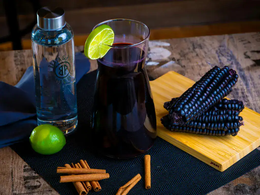 Traditional Bolivian purple api drink served with black corn, cinnamon, and lime on a wooden table.