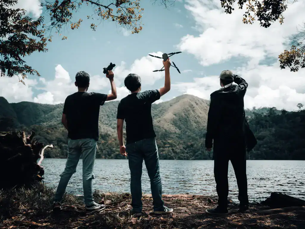 Three men holding drones by a mountain lake, silhouette against a scenic backdrop.